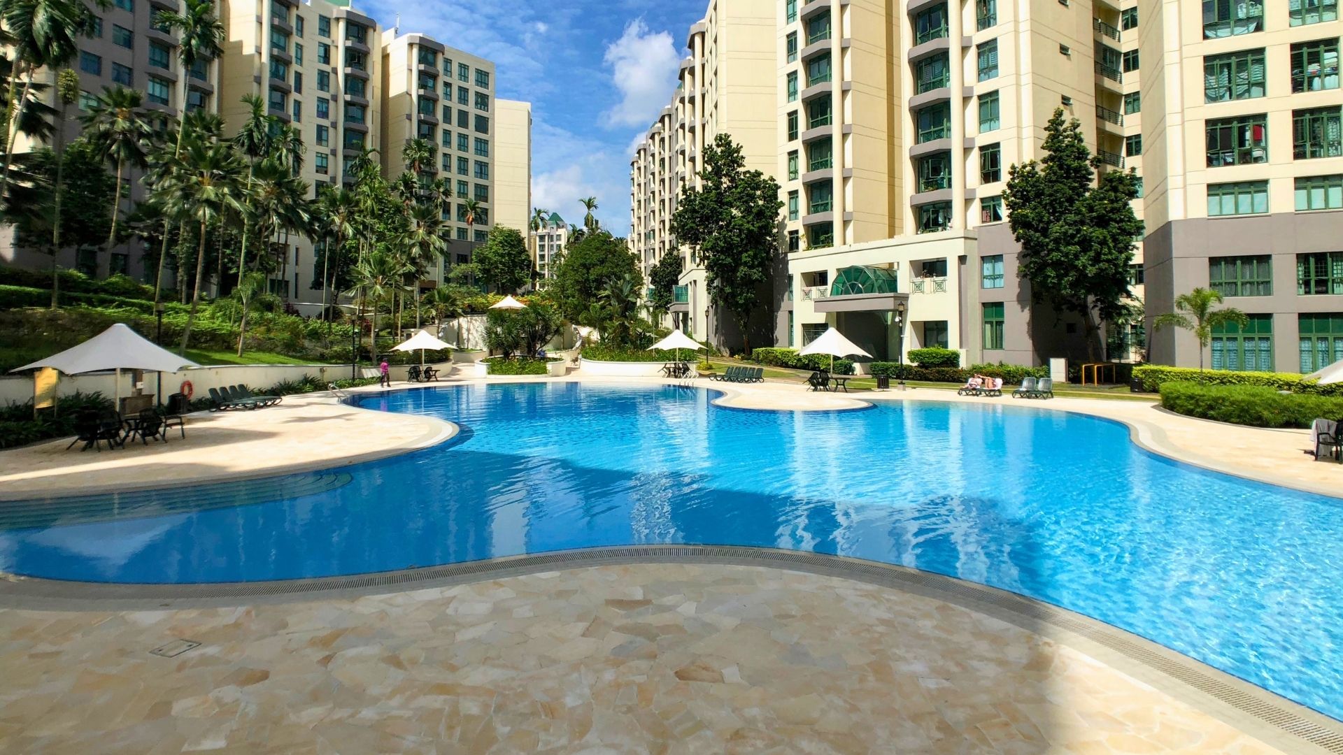 The swimming pool at Signature Park, Singapore. The edges are highly curvaceous, with clusters of shaded tables and chairs at the wide bends of the pool deck. Several condominium buildings overlook the pool.