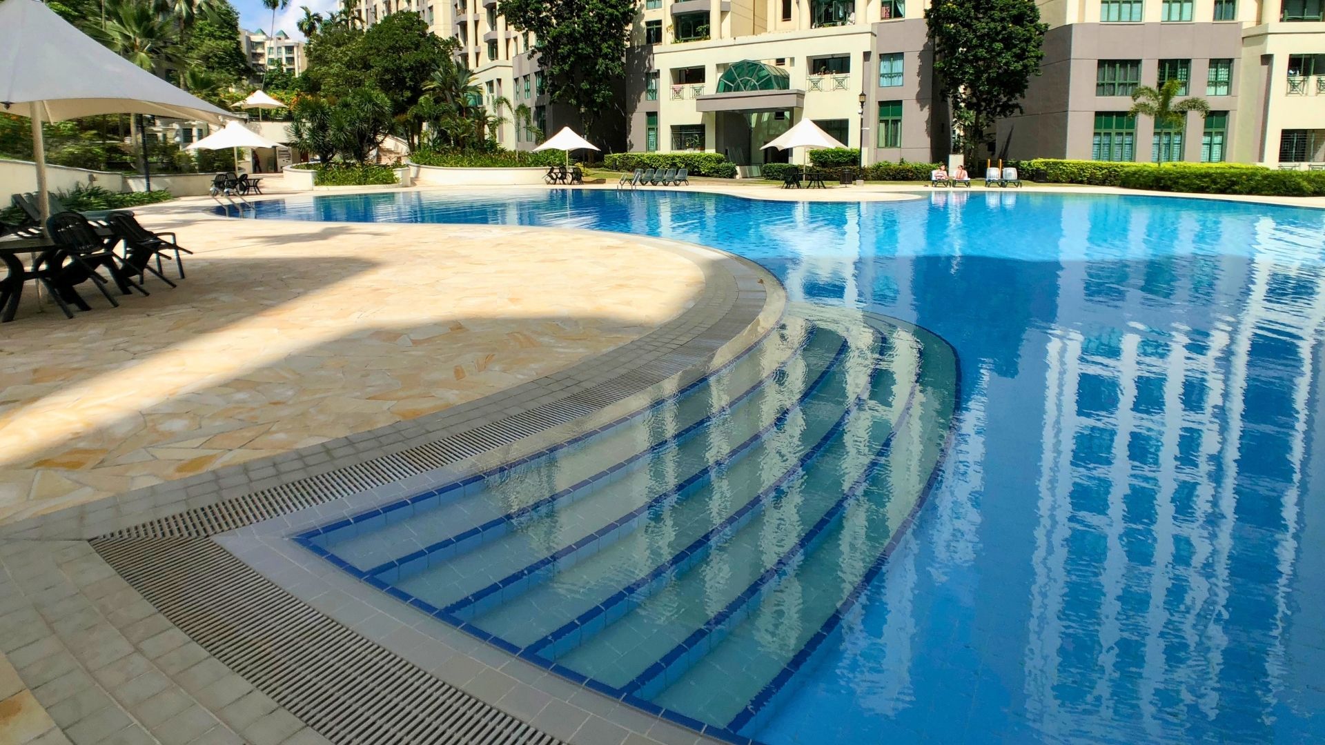 The swimming pool steps at Signature Park condominium in Singapore. They follow the curved shape of the pool, and disappear into the background. Chairs have been propped against tables under a big white umbrella in clusters around the pool deck.