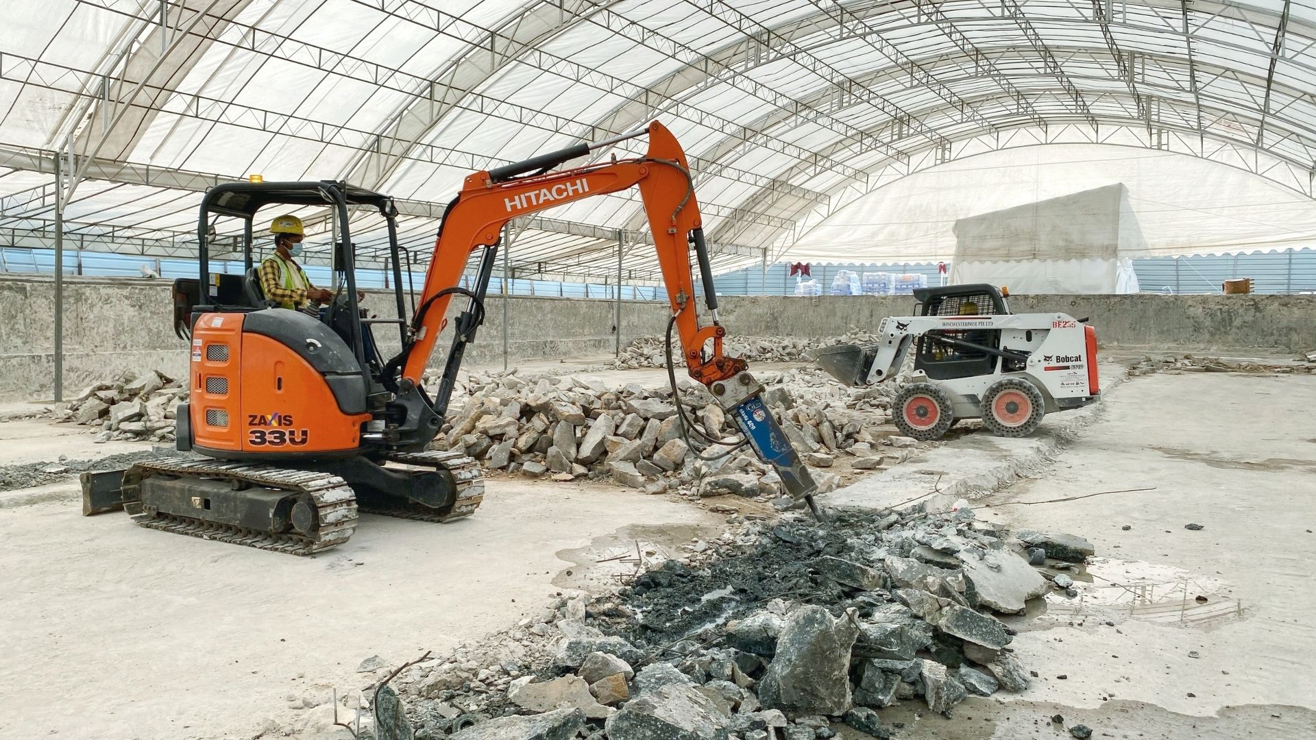 A construction worker in bright yellow safety gear operates a Hitachi swimming pool excavator at the Singapore Island Country Club. The concrete flooring is being broken up for ease of disposal.