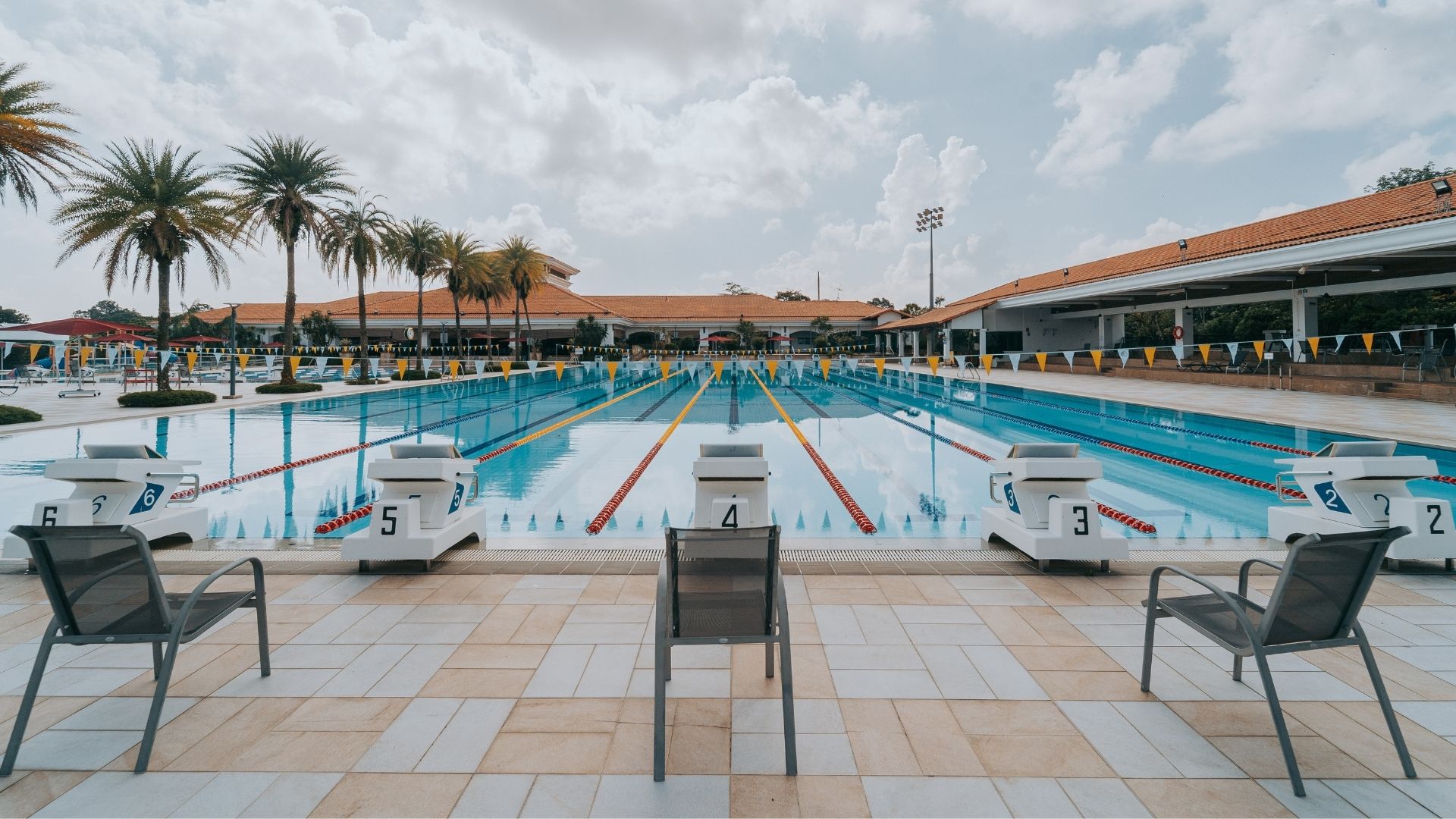 Numbered starting blocks face forward at Singapore Island Country Club's swimming pool. Pool pennants in white and yellow hang across the pool. Pool deck chairs lined neatly behind the starting blocks.