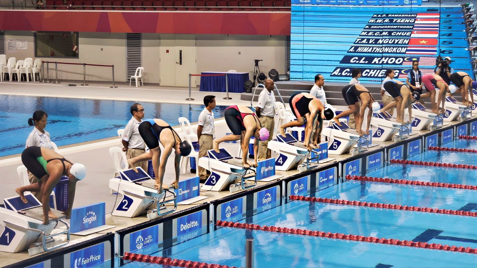 A swim competition is about to begin, as female swimmers are poised to launch off the starting blocks at the OCBC Aquatic Centre, with judges standing behind them. A digital video board is lit up to the side, featuring their names and country flags.