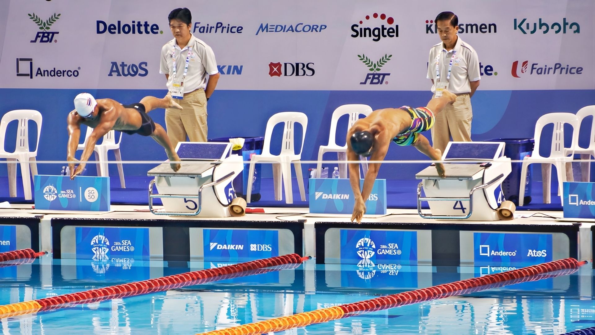 Two male swimmers are captured in mid-air, having just launched off the starting blocks at the OCBC Aquatic Centre in Singapore. Competition judges stand behind them. Renown commercial brand sponsors are featured on a large board behind.