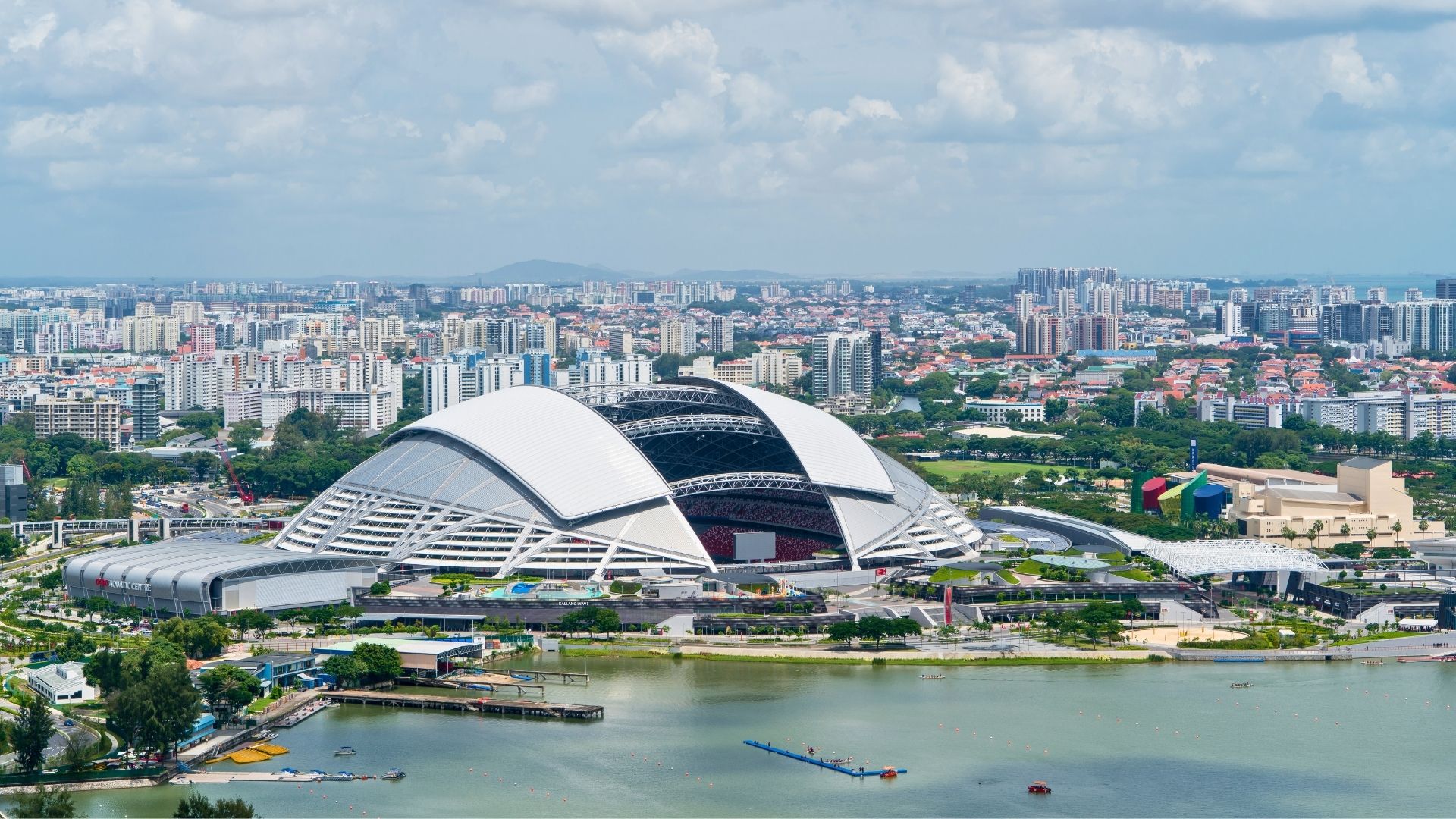 Aerial overview of the Singapore Sports Hub/The Kallang, which is in the shape of a dome. The centre of the dome has been segmented into an open-air sports venue. The Hub is surrounded by city structures and encompassed by Kallang Basin.
