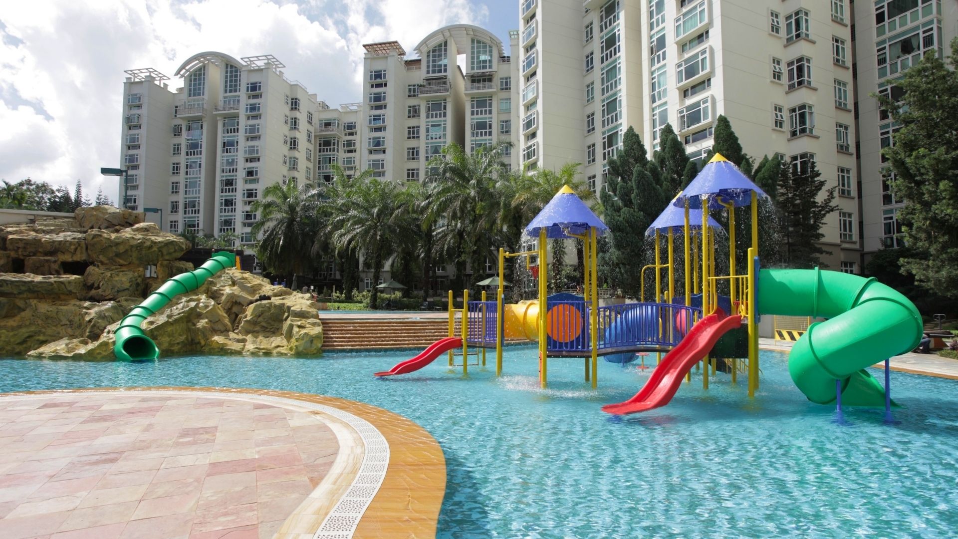 The newly-renovated children’s pools at Summerhill Condominium. A full-featured wet playground occupies the centre, and a bright green tube slide sits embedded within rock landscaping behind it.