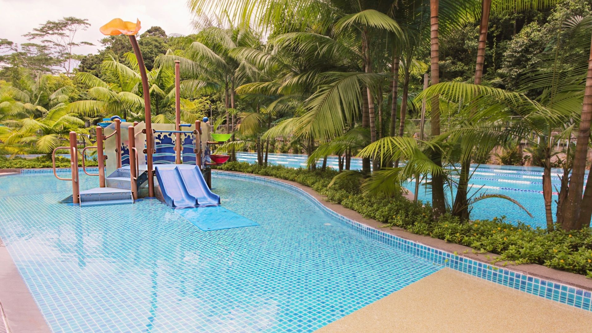The upgraded children's pool and colourful wet playground at Temasek Club in Singapore, complete with slides, grab rails, and a floral structure curved upwards. The main lap pool is behind it, separated by a row of bushes and trees.