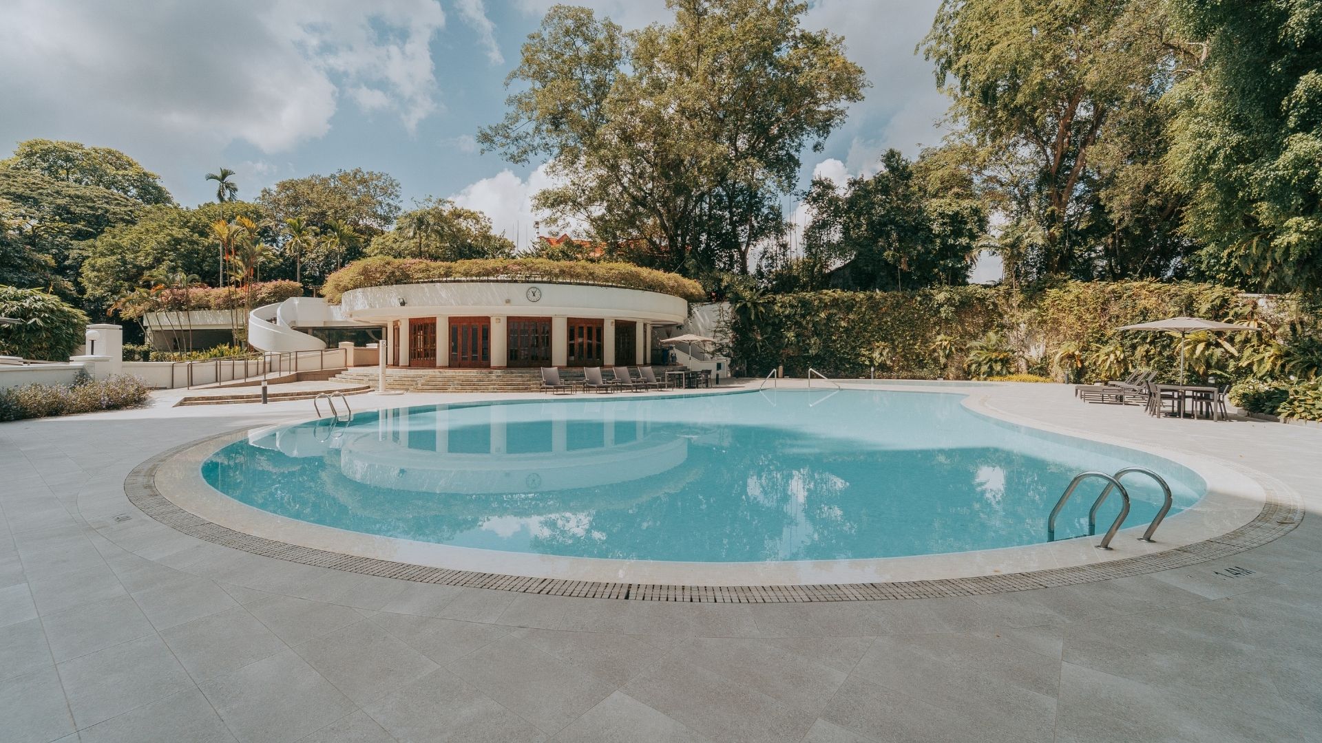 The swimming pool at The Arcadia after renovation, featuring a Spanish porcelain-tiled pool deck in neutral tones, with matching porcelain tile gratings. A short building with an analog clock shows the time in the background.