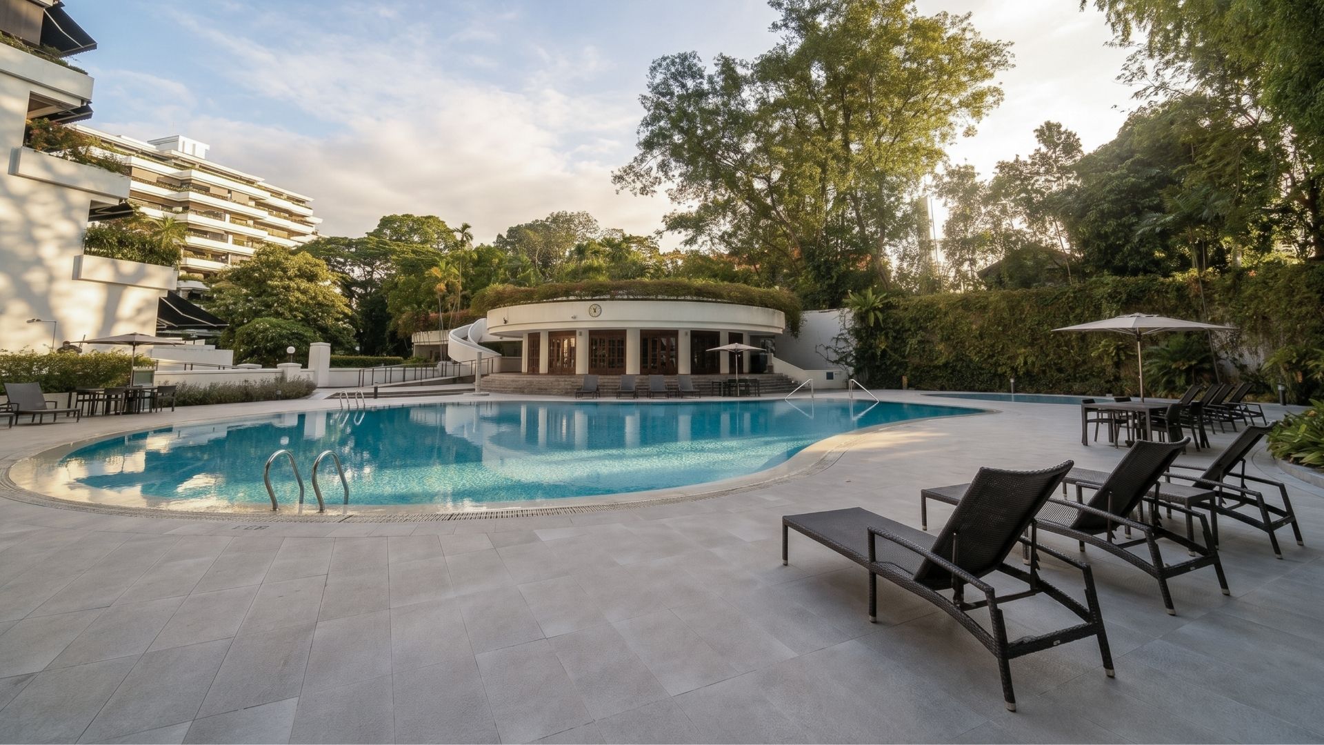 The renovated pool deck at The Arcadia Condominium in Singapore, with adjustable lounge chairs and shaded table areas. It features a Spanish porcelain-tiled deck, and matching porcelain tile gratings. Grab rails are found around the pool.
