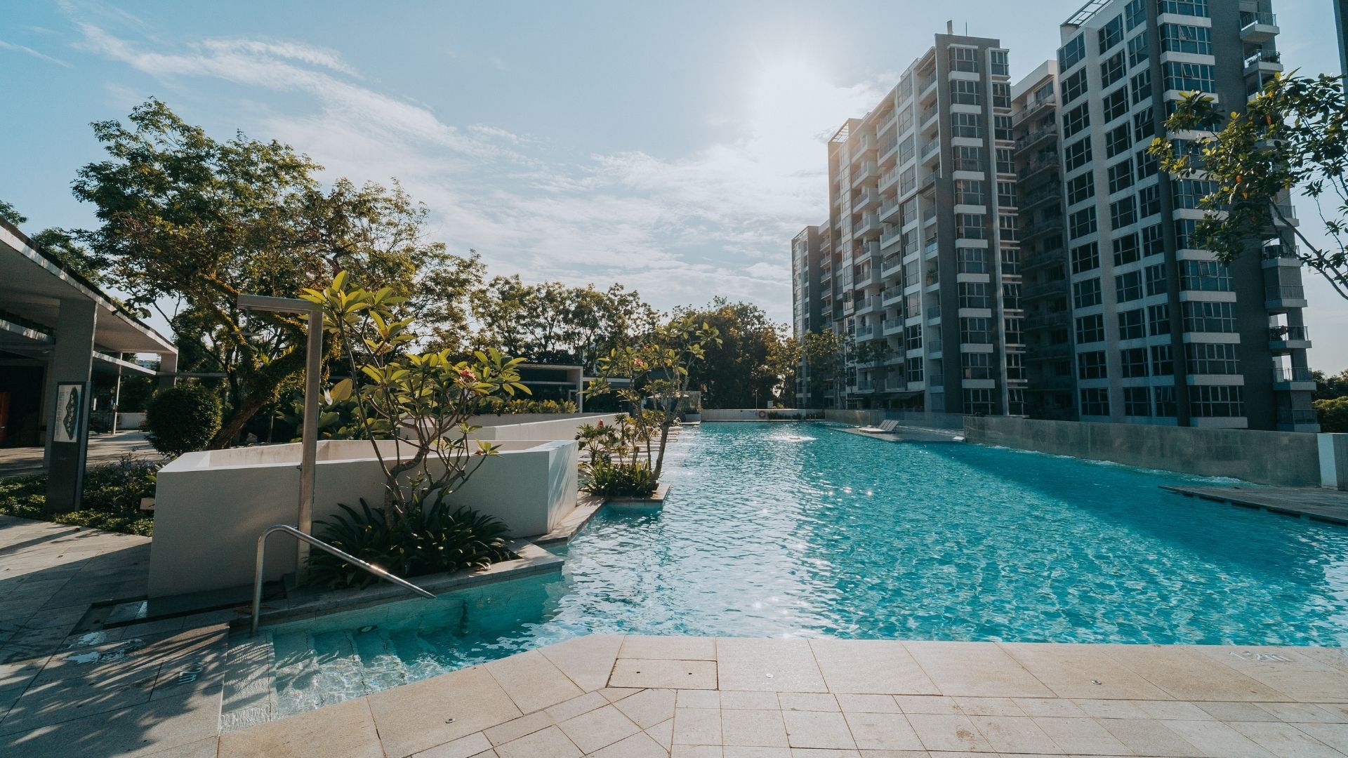 The renovated concrete swimming pool at The Estuary Condominium in Singapore, featuring well-laid deck tiles, and carefully crafted steps leading into the water with a grab rail. The pool is long, and spans across several residential blocks.