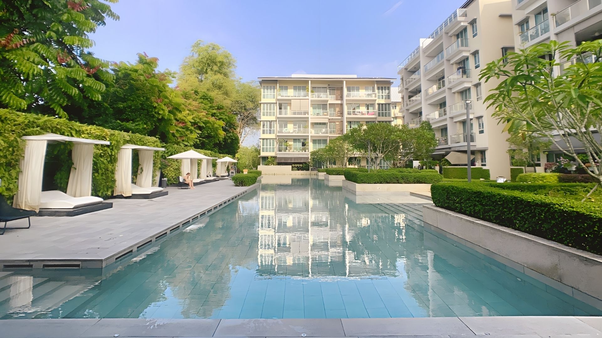 Ground-level view of the 50-metre swimming pool at The Glyndebourne Condominium in Singapore. Resort-style cabanas draped with breezy white curtains and deck loungers are spaced out evenly amidst manicured landscaping.