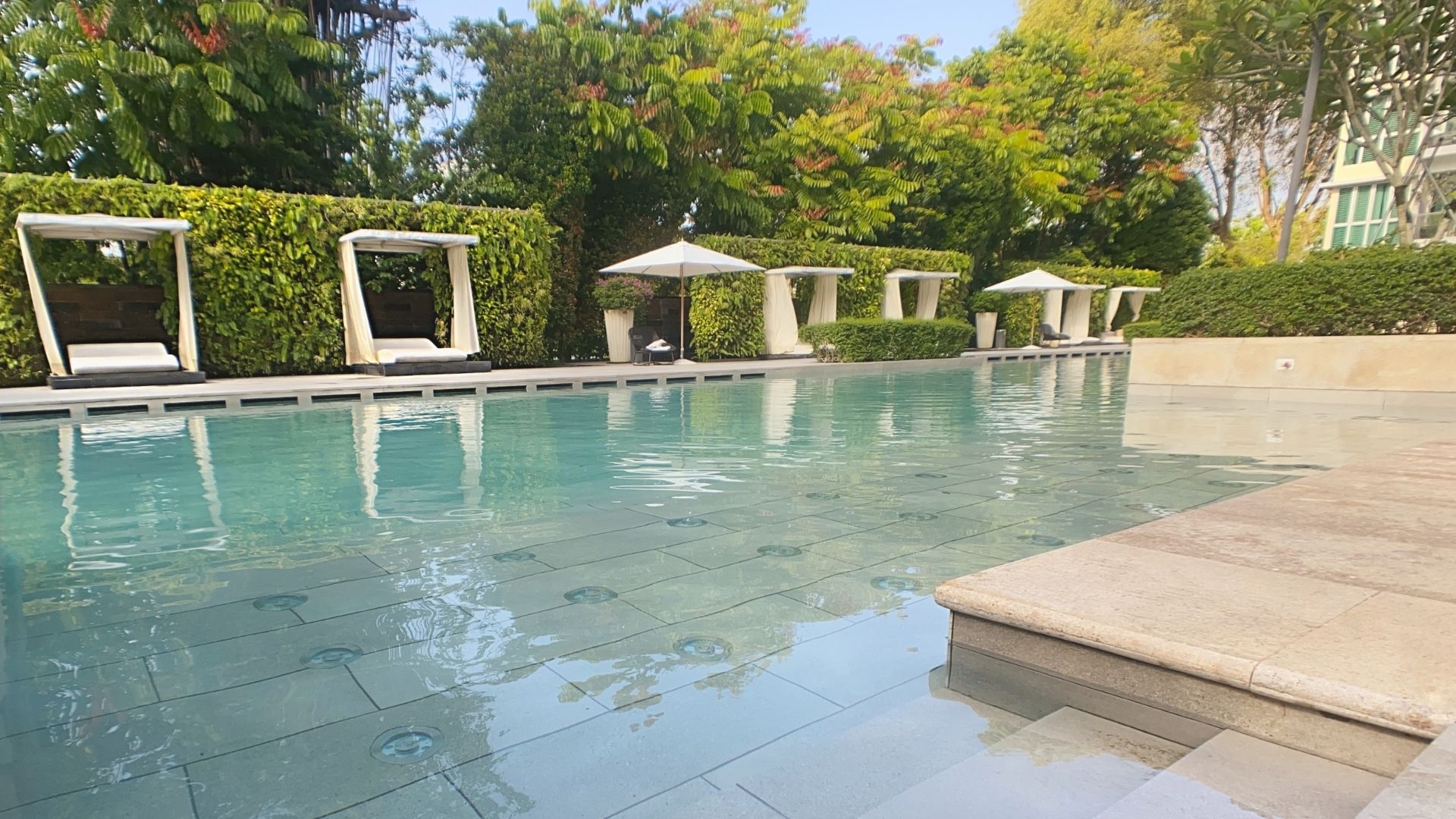 View across the swimming pool at the The Glyndebourne Condominium, facing towards the pool deck with comfortable cabanas and lounge chairs. Wide pool steps in Spanish large-format tiles lead into the pool, with underwater lights on the pool floor.