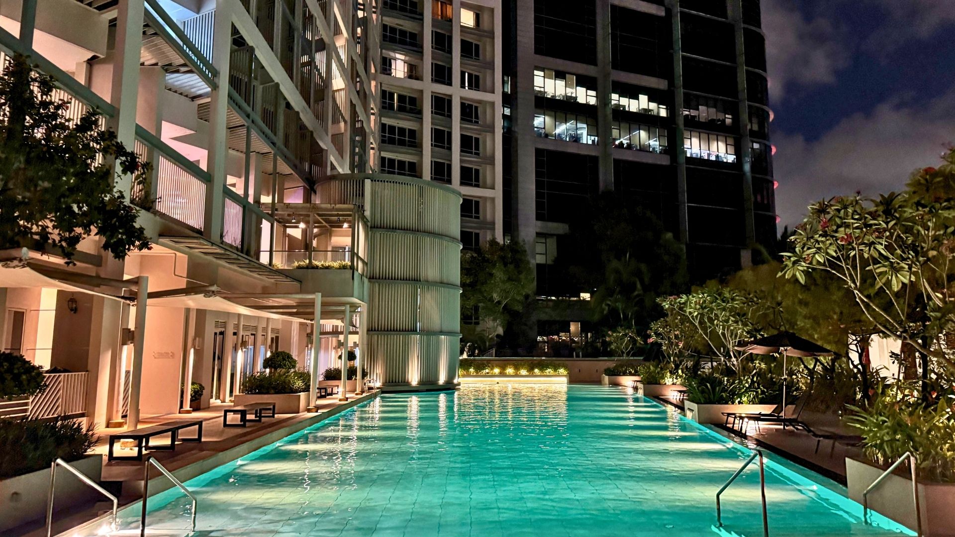 The upgraded swimming pool at Visioncrest Residence in Singapore at night. Underwater lights and ambient lighting from the pool deck light the pool up brightly. Steps lead into the pool at the left and right corners, with hand rails for support.