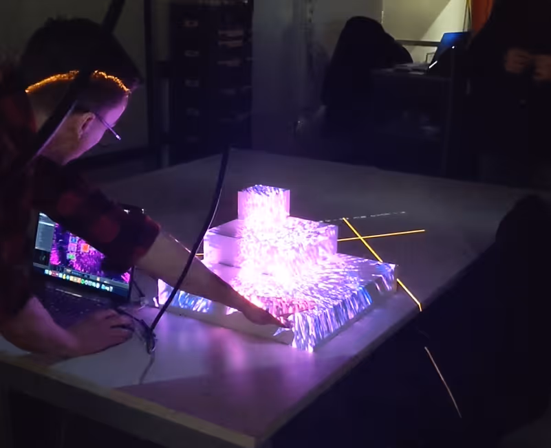 Person interacting with a laptop and a 3D stepped model projecting colorful, dynamic light patterns on a table.
