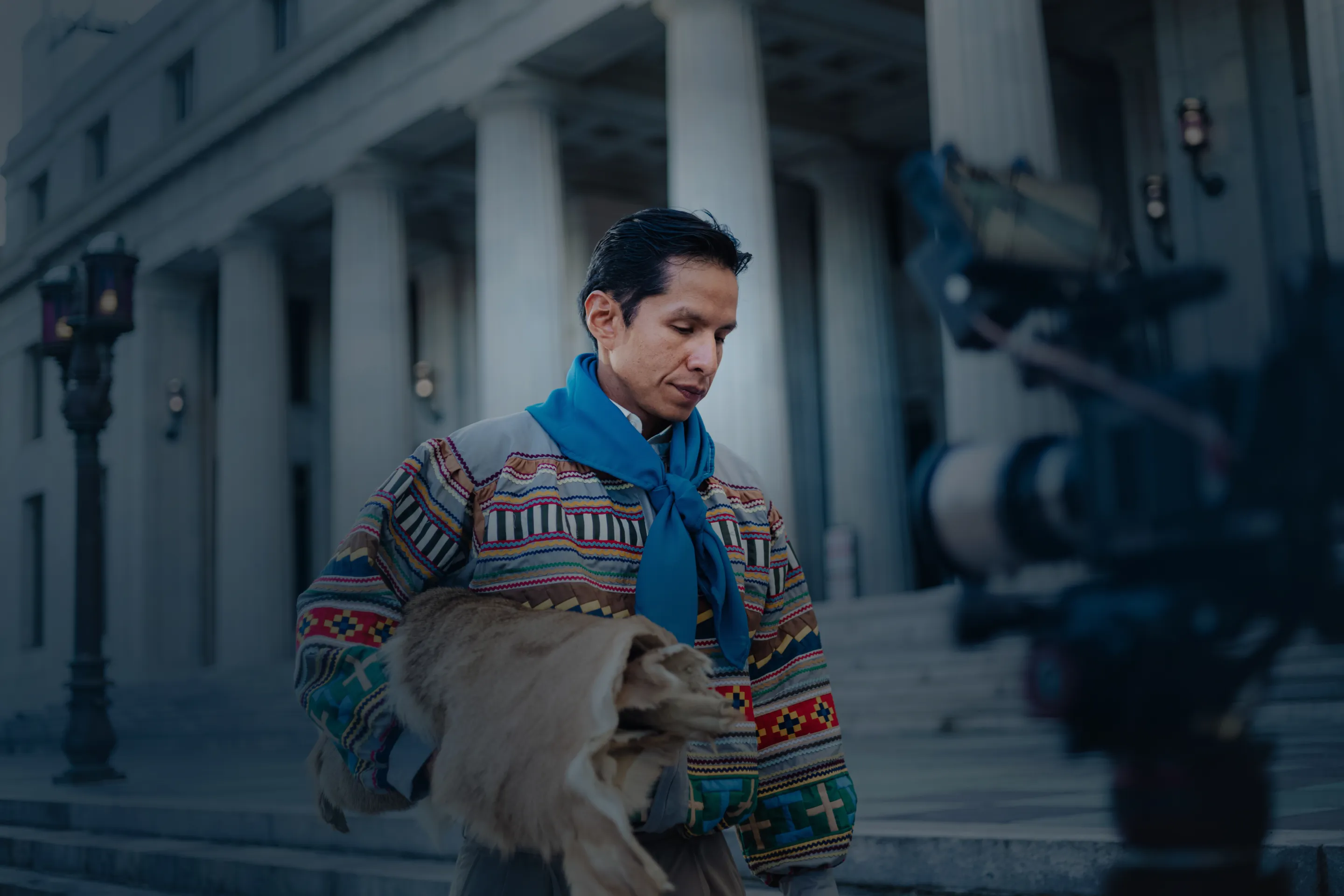 Man wearing a colorful patterned jacket and blue scarf holding a fur garment in front of a building with columns.