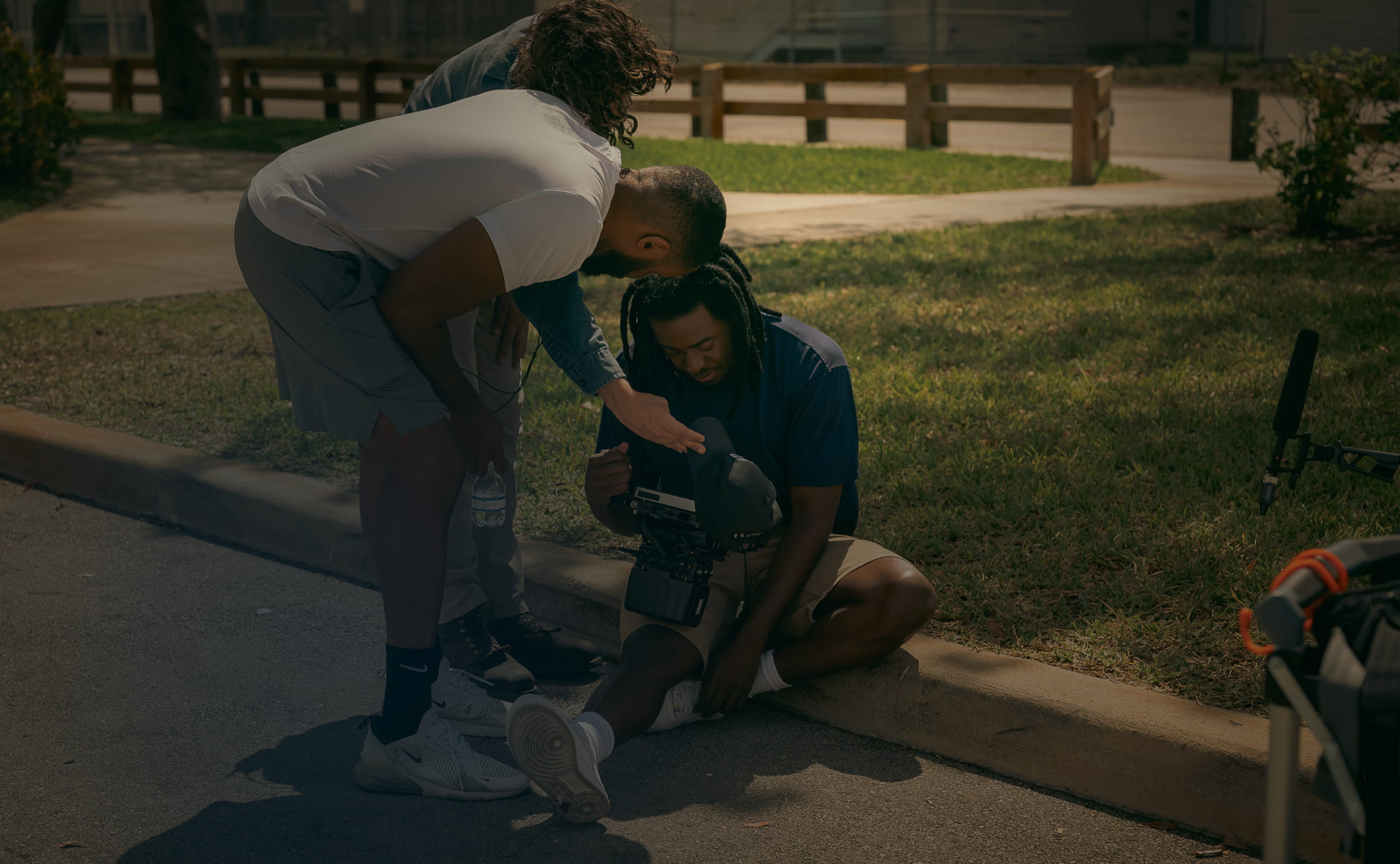 Three men examining a camera while one sits on a curb and others lean over him on a sunny day.