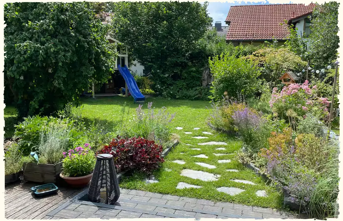 Blick von der Terrasse in ein Gartengrundstück mit Polygonsteinen als Trittplatten und zwei großen, blühenden, organisch geformten Beeten.