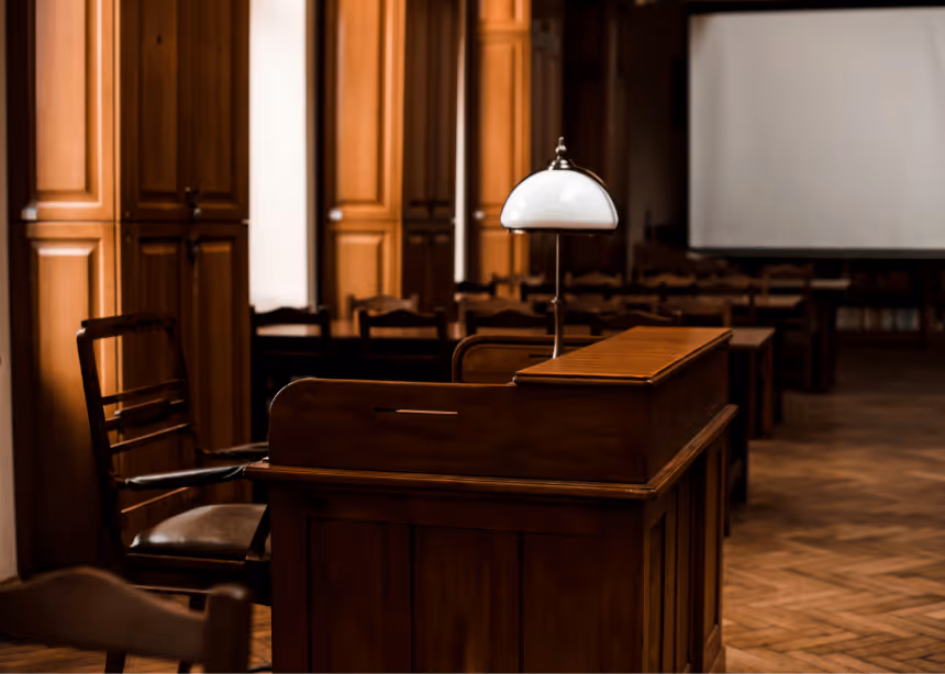 Salle de tribunal avec un bureau en bois et une lampe blanche, chaises en bois et un grand écran blanc en arrière-plan.