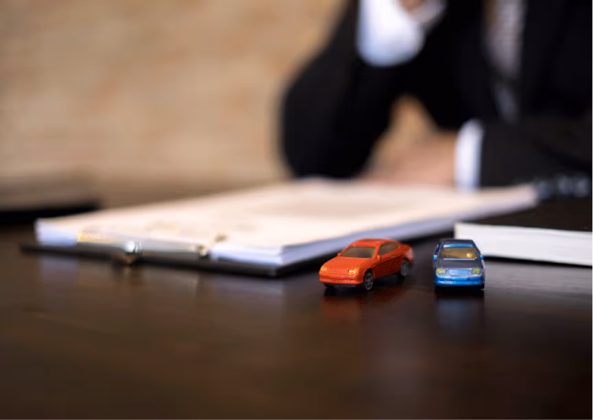 Deux petites voitures jouets rouges et bleues posées sur une table devant un clipboard avec des papiers et une personne floutée en arrière-plan.