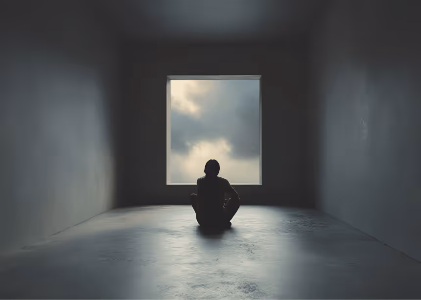 Silhouette d'une personne assise seule sur le sol dans une pièce sombre face à une fenêtre carrée montrant un ciel nuageux.