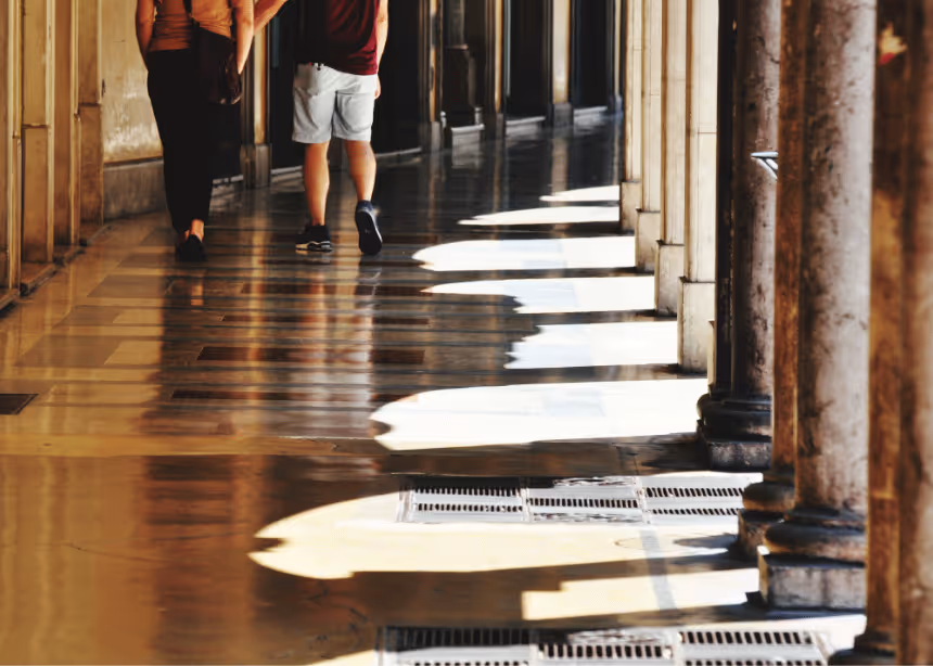 Deux personnes marchant dans un couloir bordé de colonnes avec lumière et ombres au sol poli.