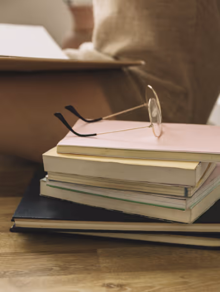 Une pile de livres et de carnets posée sur un sol en bois avec une paire de lunettes rondes posée dessus, et une personne assise en arrière-plan flouté.