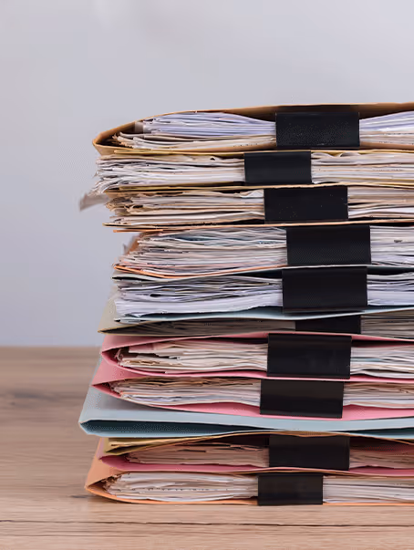 Pile de dossiers multicolores épais, attachés avec des grandes pinces noires, posée sur un bureau en bois.