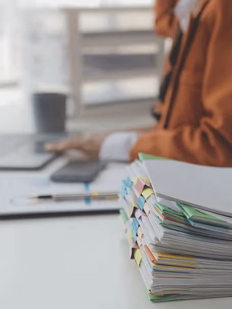 Pile de dossiers et papiers avec plusieurs onglets colorés sur une table de bureau, une personne en arrière-plan portant une veste marron utilise une tablette.