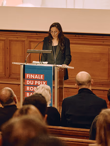 Une femme portant des lunettes parle à un pupitre en bois devant un public assis, lors de la finale du Prix Rob des Bains.