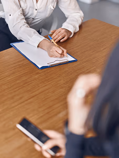 Une personne écrivant sur une feuille blanche fixée à un clipboard, tandis qu'une autre personne en arrière-plan tient un smartphone.