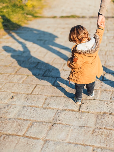 Un enfant portant une veste marron tient la main d'un adulte sur un chemin pavé, avec leurs ombres longues projetées au sol.