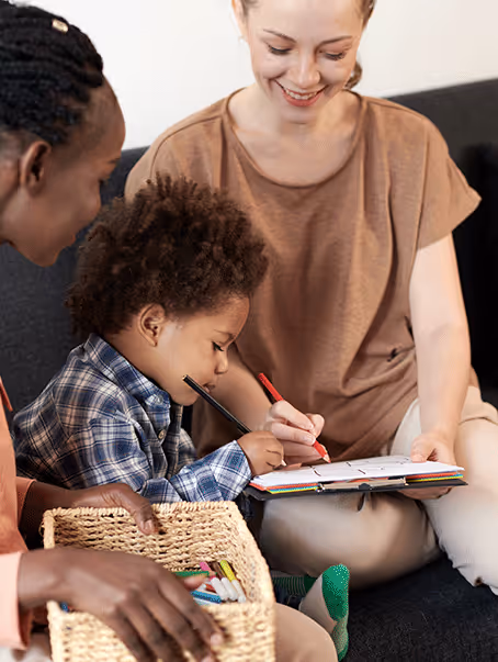 Un enfant dessine avec un crayon tandis qu'une femme souriante le guide et un autre adulte tient un panier de crayons.