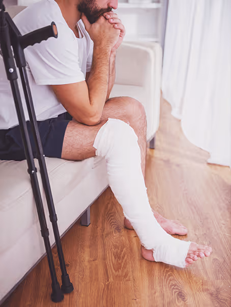 Homme assis sur un canapé avec une jambe en plâtre et des béquilles appuyées à côté.
