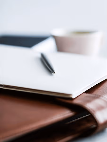 Stylo posé sur un carnet blanc ouvert, avec un porte-documents en cuir marron en dessous et une tasse floue en arrière-plan.