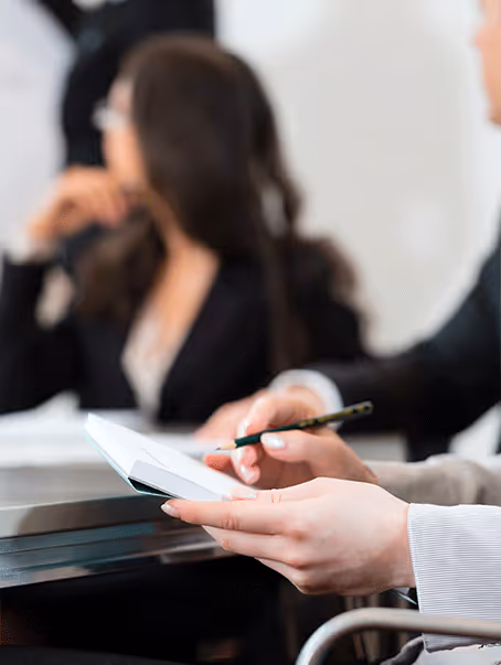 Personne prenant des notes sur un carnet lors d'une réunion professionnelle avec d'autres participants flous en arrière-plan.