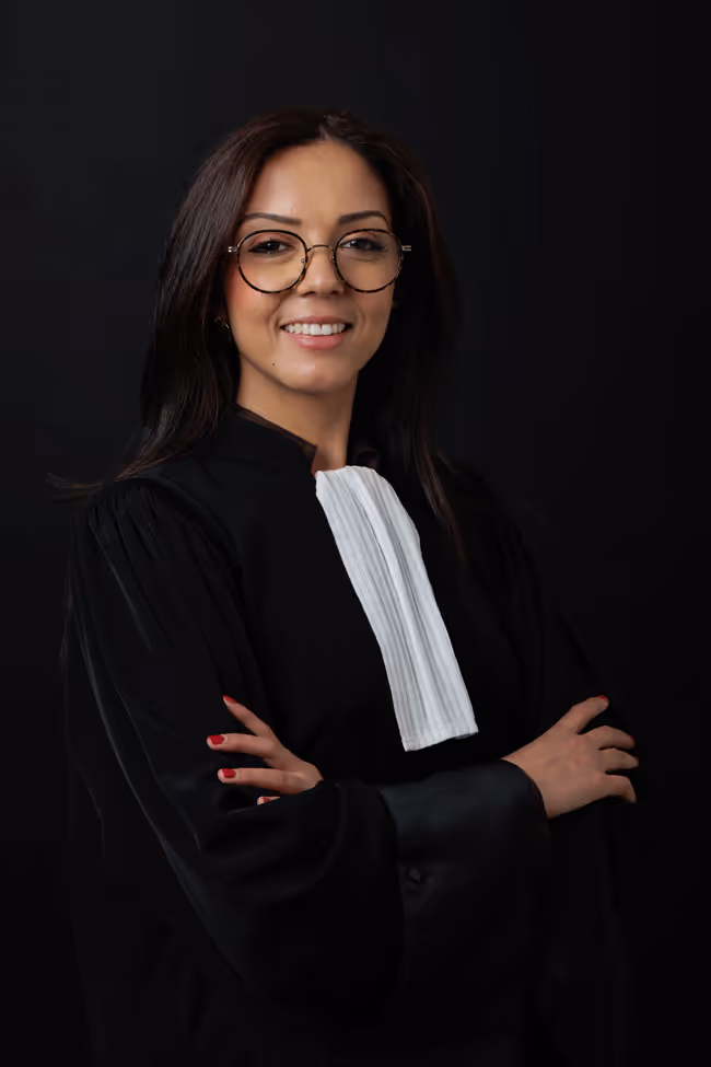 Portrait d'une femme avocate souriante portant une robe noire et une bavette blanche avec des lunettes rondes, bras croisés devant un fond noir.