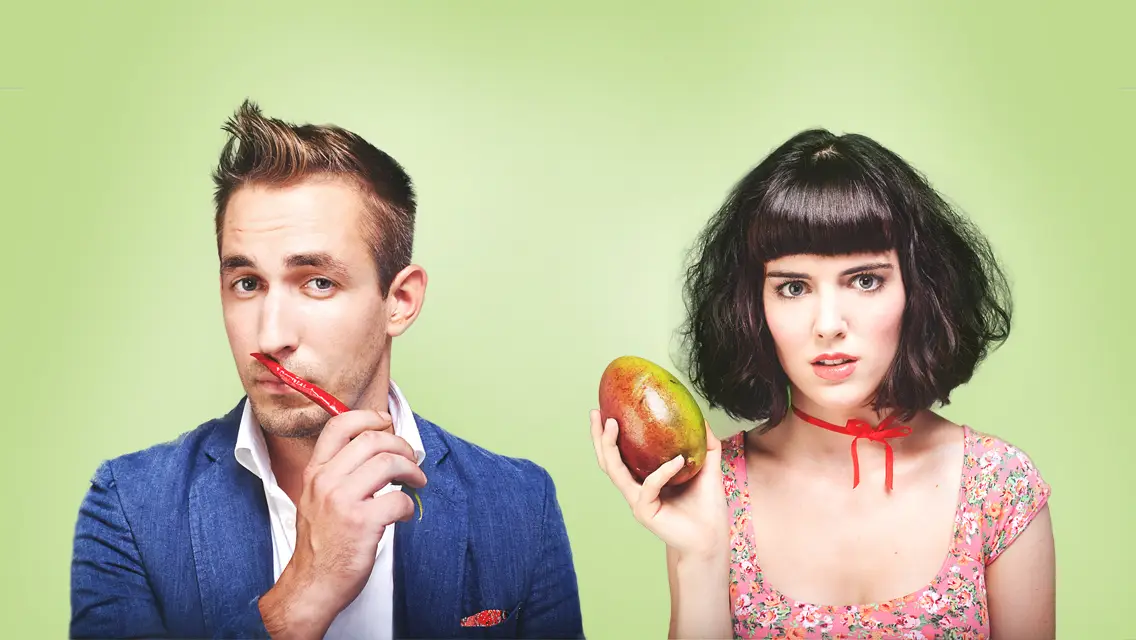 Man in blue jacket holding red chili pepper near his mouth and woman in floral dress holding a mango, both against a green background.