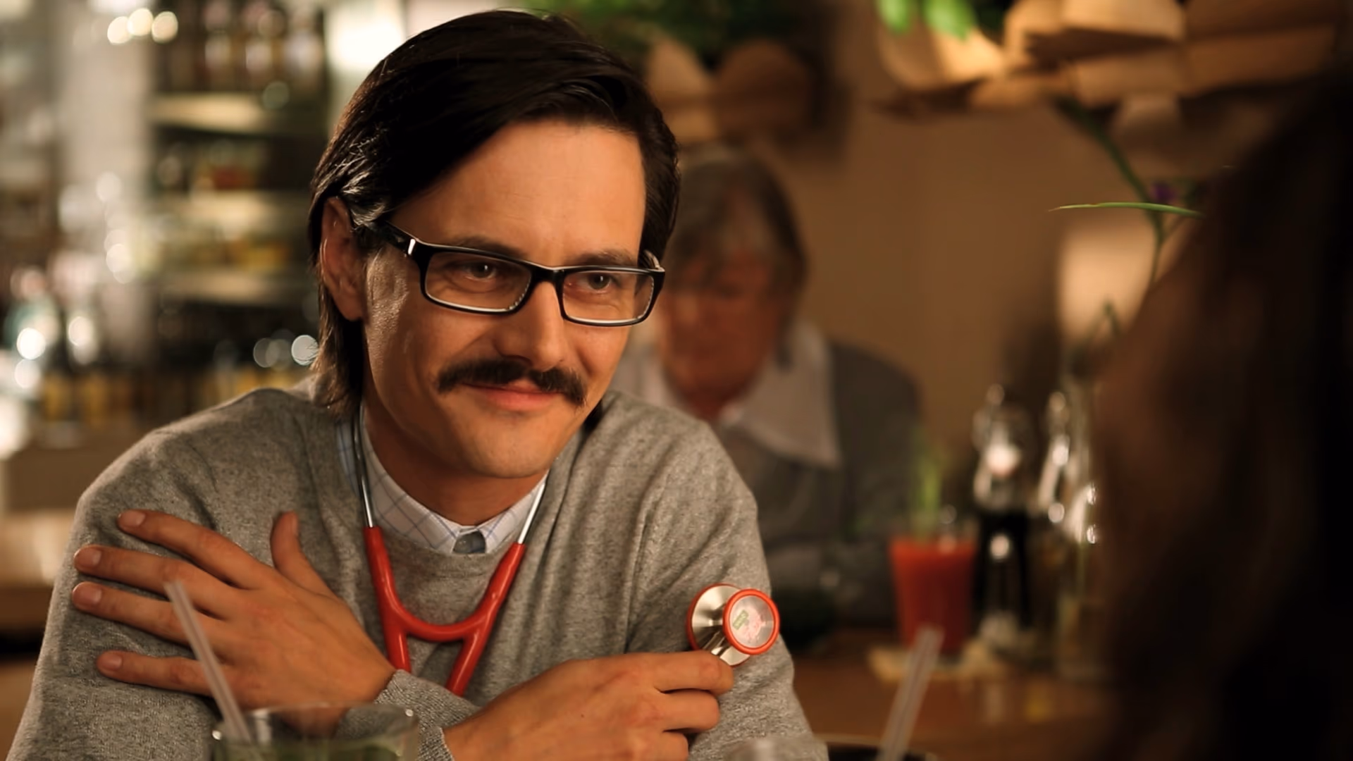 Man with a mustache, glasses, and a stethoscope around his neck sitting at a table in a cozy setting.