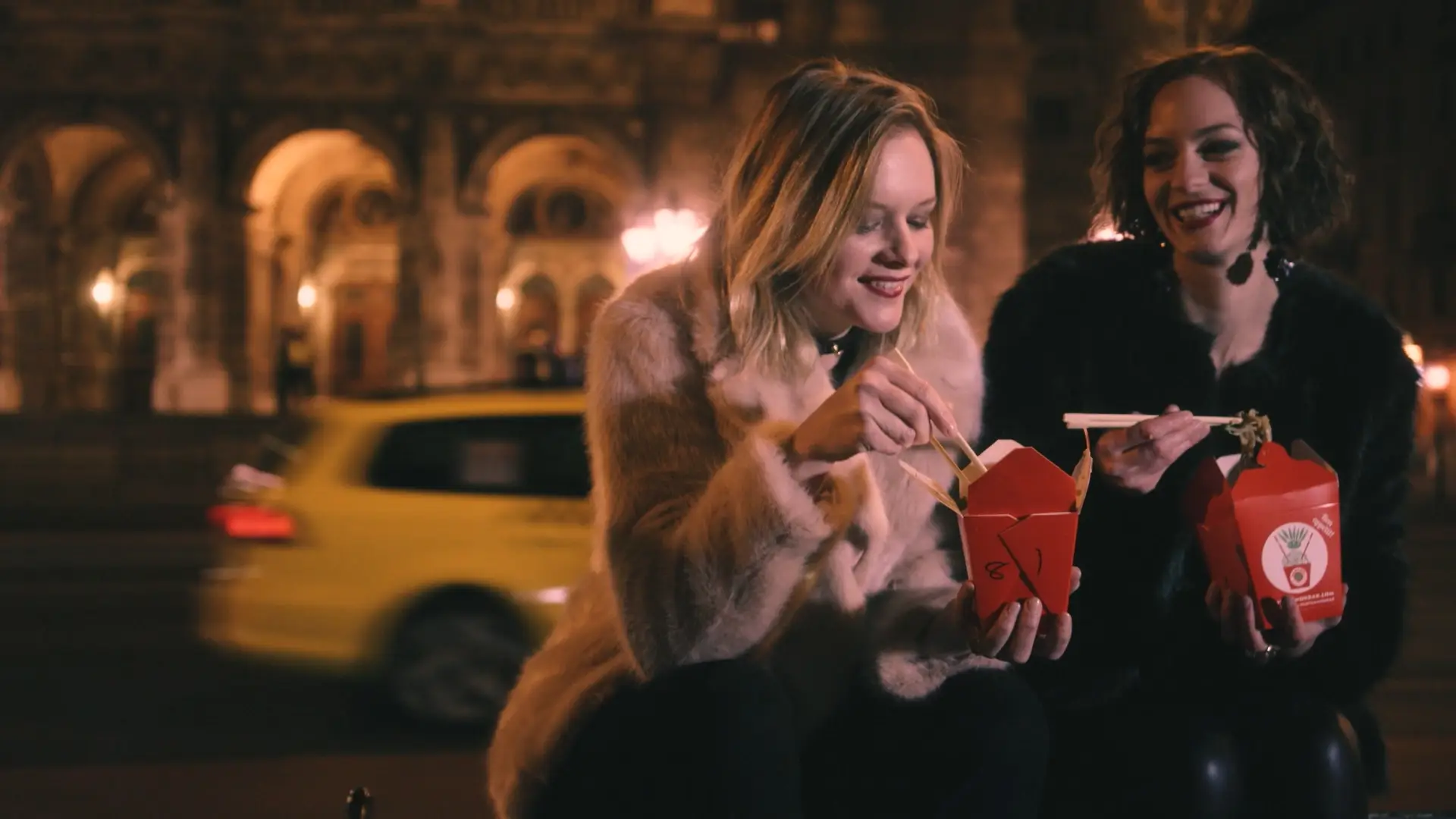 Two women in fur coats eating Asian takeout with chopsticks outdoors at night with a yellow car and lit arches in the background.