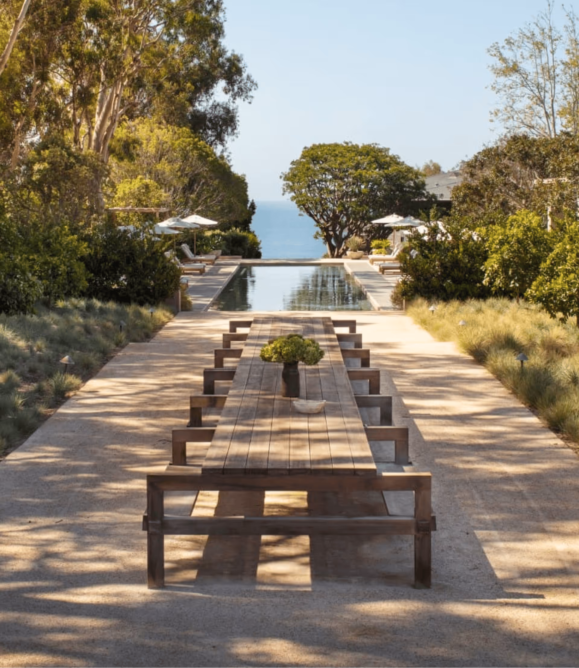 Long wooden outdoor dining table with benches by a pool surrounded by trees and ocean view.
