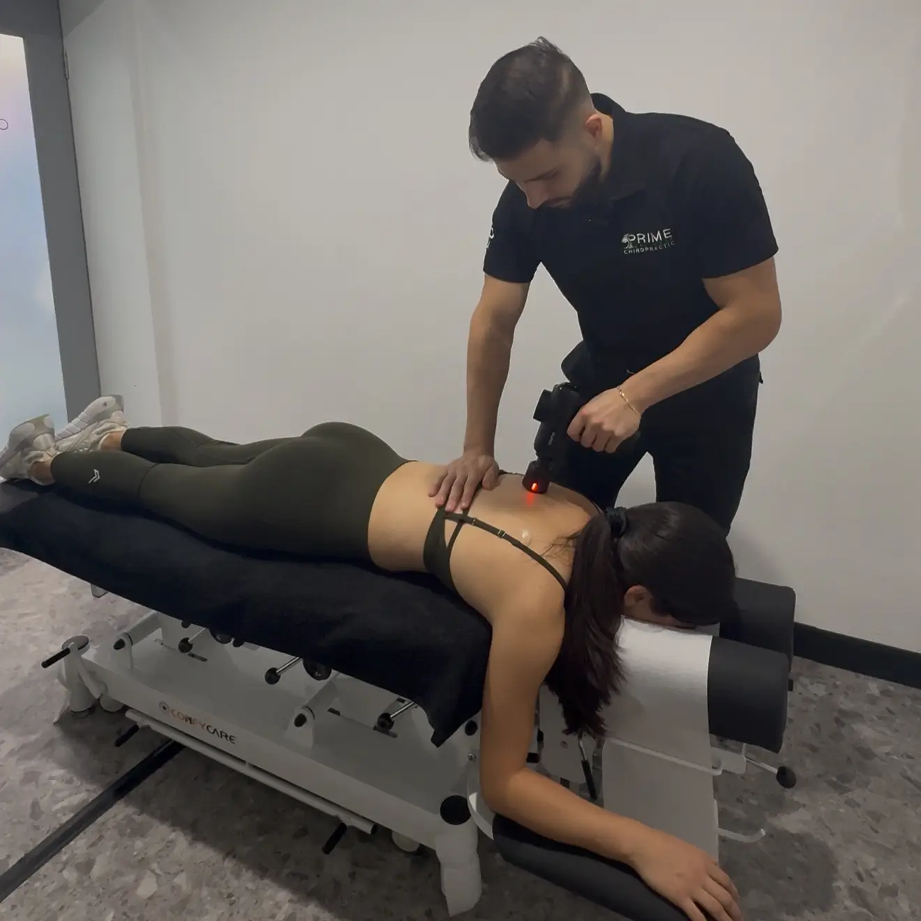 A chiropractor wearing a black shirt with 'Prime Chiropractic' on it uses a handheld device emitting a red light on the upper back of a woman lying face down on a treatment table in a clinical room.