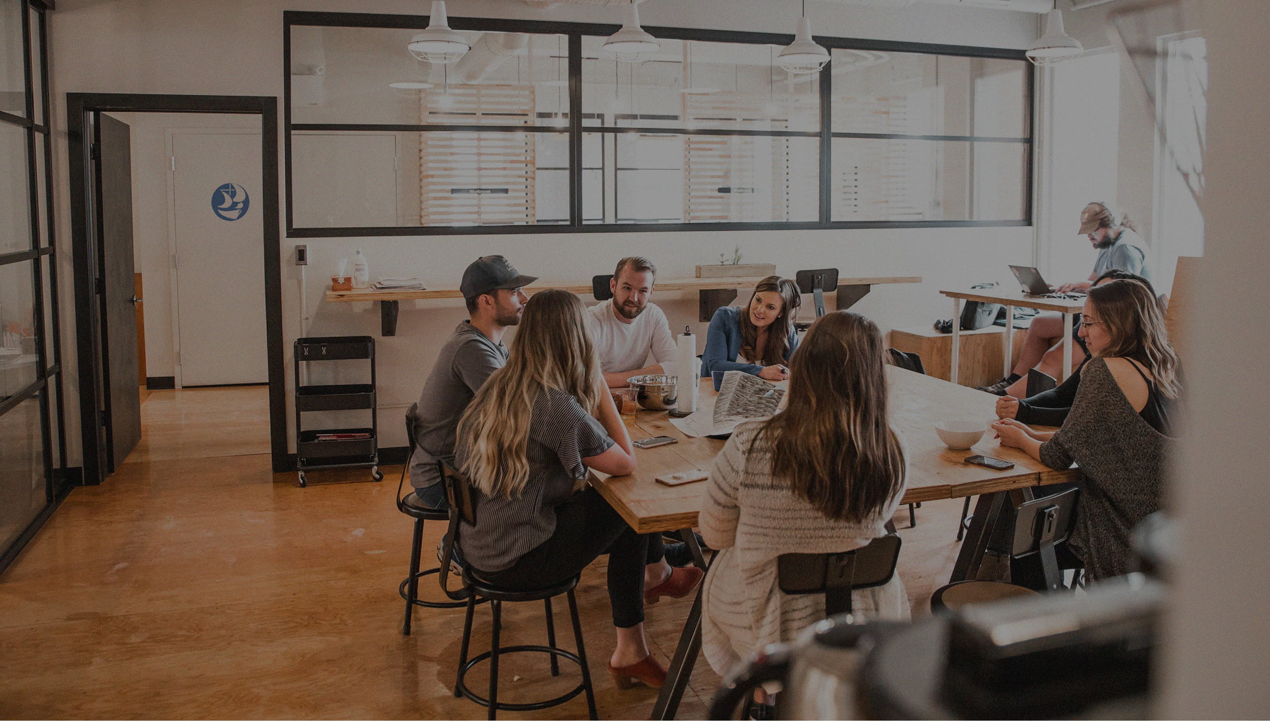 Seven people engaged in discussion around a wooden table in a modern office space with large windows and wooden floors.
