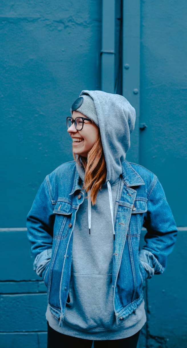 Young woman smiling and looking to the side, wearing glasses, a gray beanie, gray hoodie, and denim jacket against a blue wall.