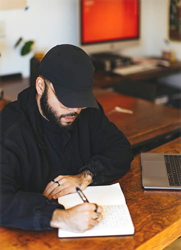 Man wearing a black cap and jacket writing in a notebook at a wooden table with a laptop nearby.