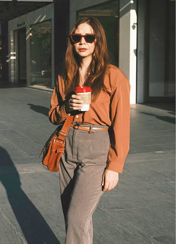 Woman wearing sunglasses, a brown blouse, and gray pants holding a takeaway coffee cup and a brown shoulder bag, standing outdoors in sunlight.