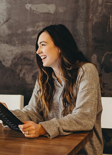 Smiling woman with long brown hair wearing a gray sweater, sitting at a wooden table and holding a black tablet.
