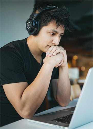 Young man wearing headphones sitting at a desk, thoughtfully looking at a laptop screen.