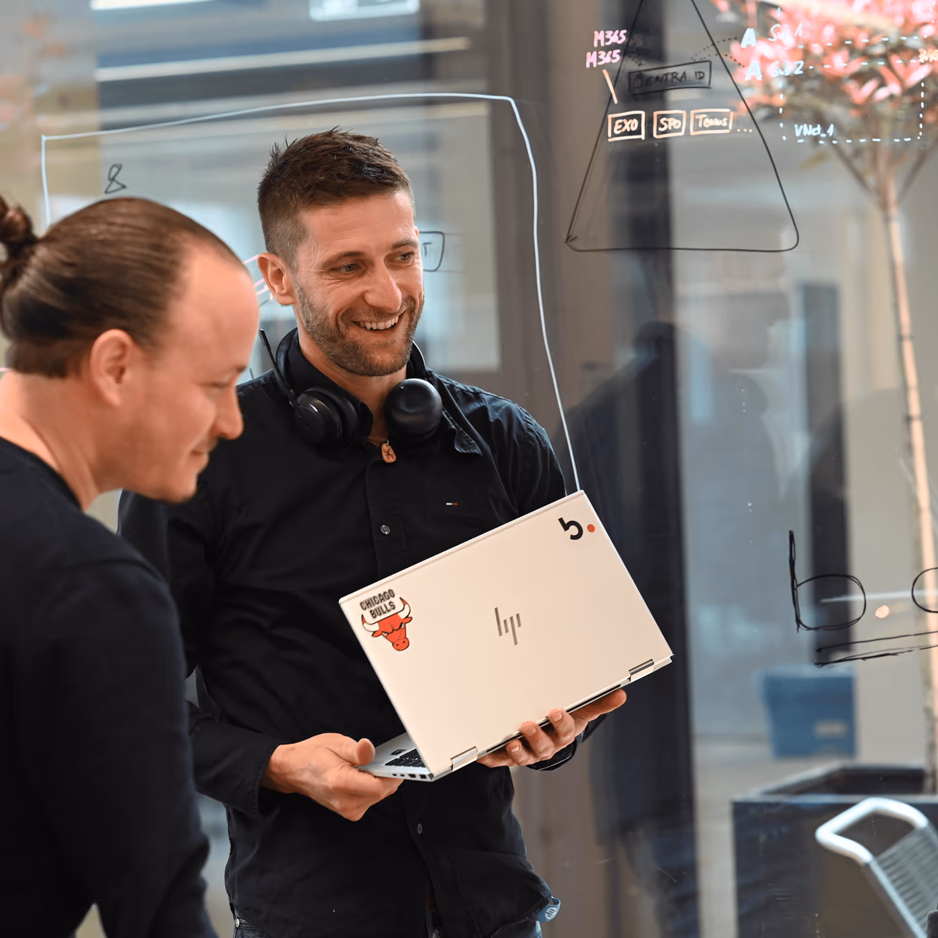 Zwei Mitarbeiter betrachten gemeinsam einen Laptop vor einer beschriebenen Glaswand im Büro