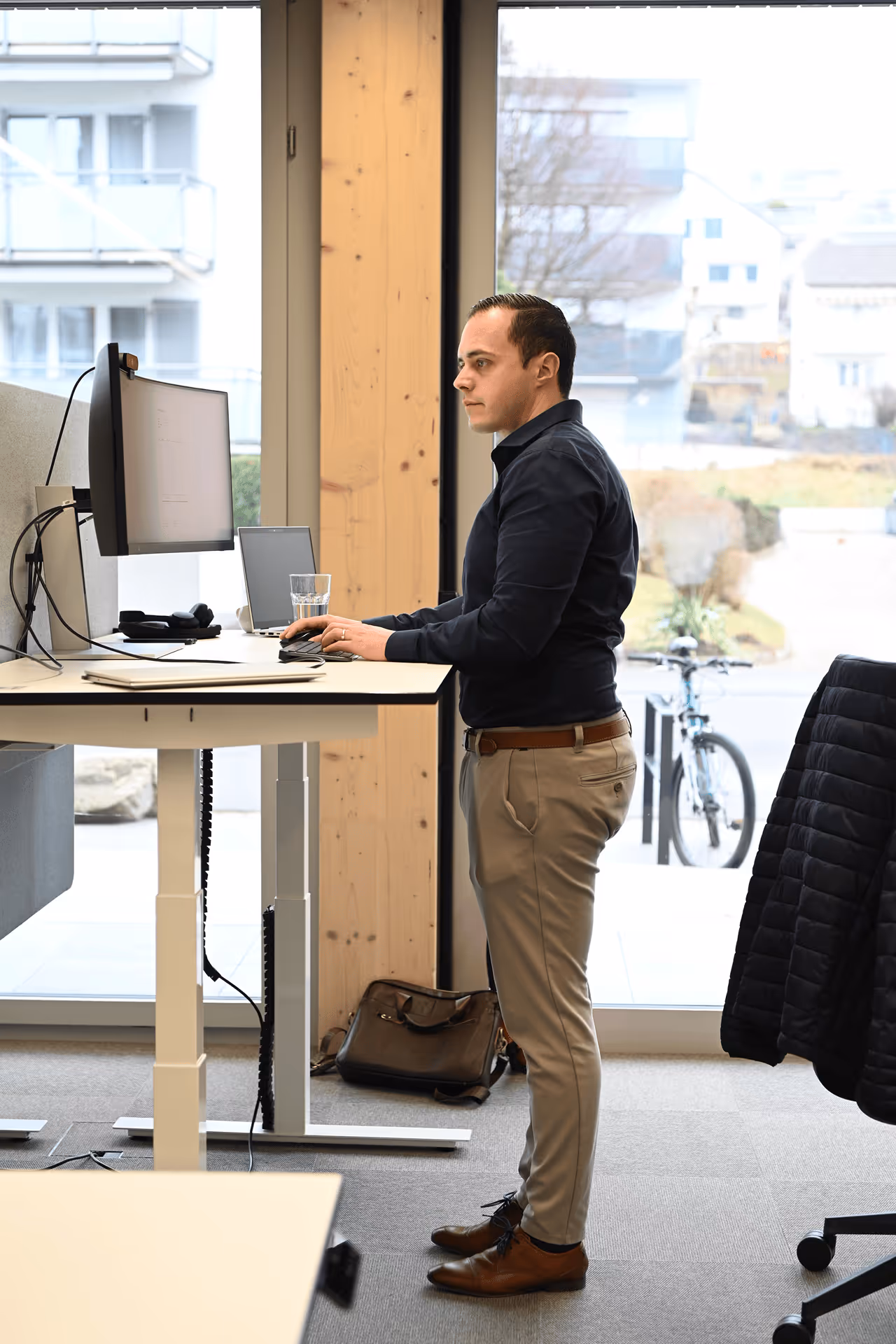 Mitarbeiter steht am höhenverstellbaren Schreibtisch und arbeitet an Laptop und Bildschirm im Büro