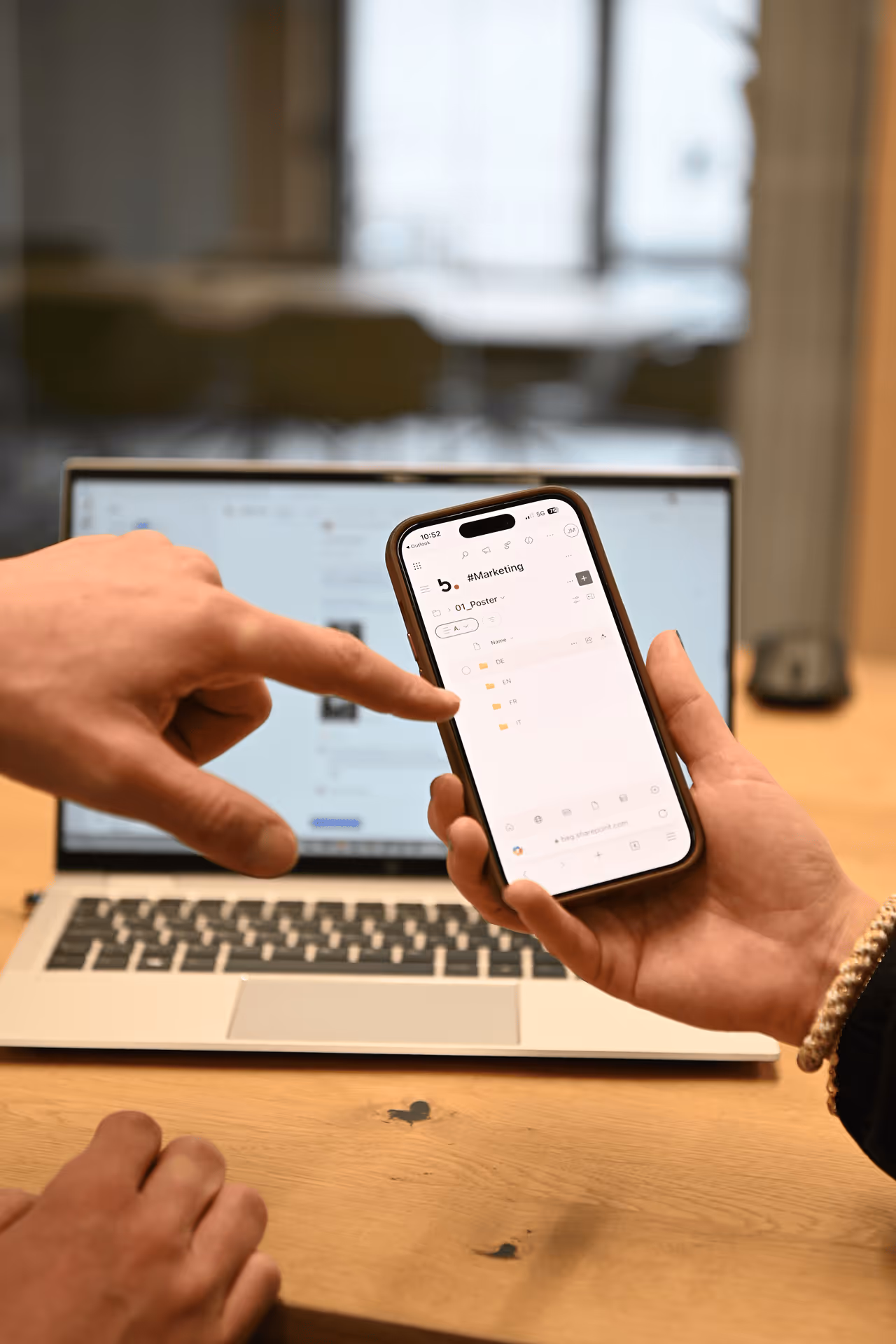 Eine Person zeigt auf das Smartphone in der Hand einer anderen, im Hintergrund ein Laptop auf dem Holztisch