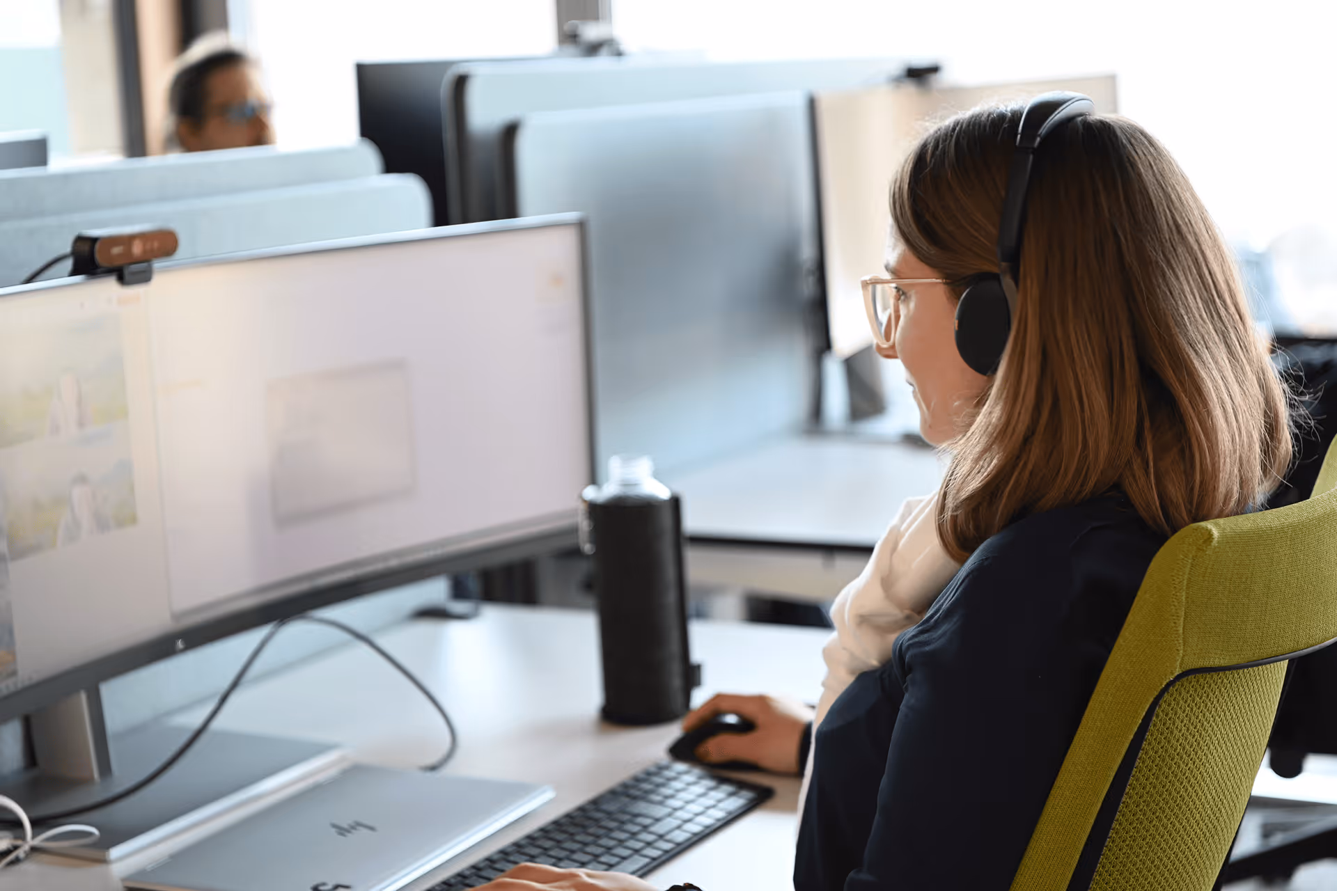 Mitarbeiterin mit Kopfhörern am Arbeitsplatz mit zwei Monitoren im Grossraumbüro