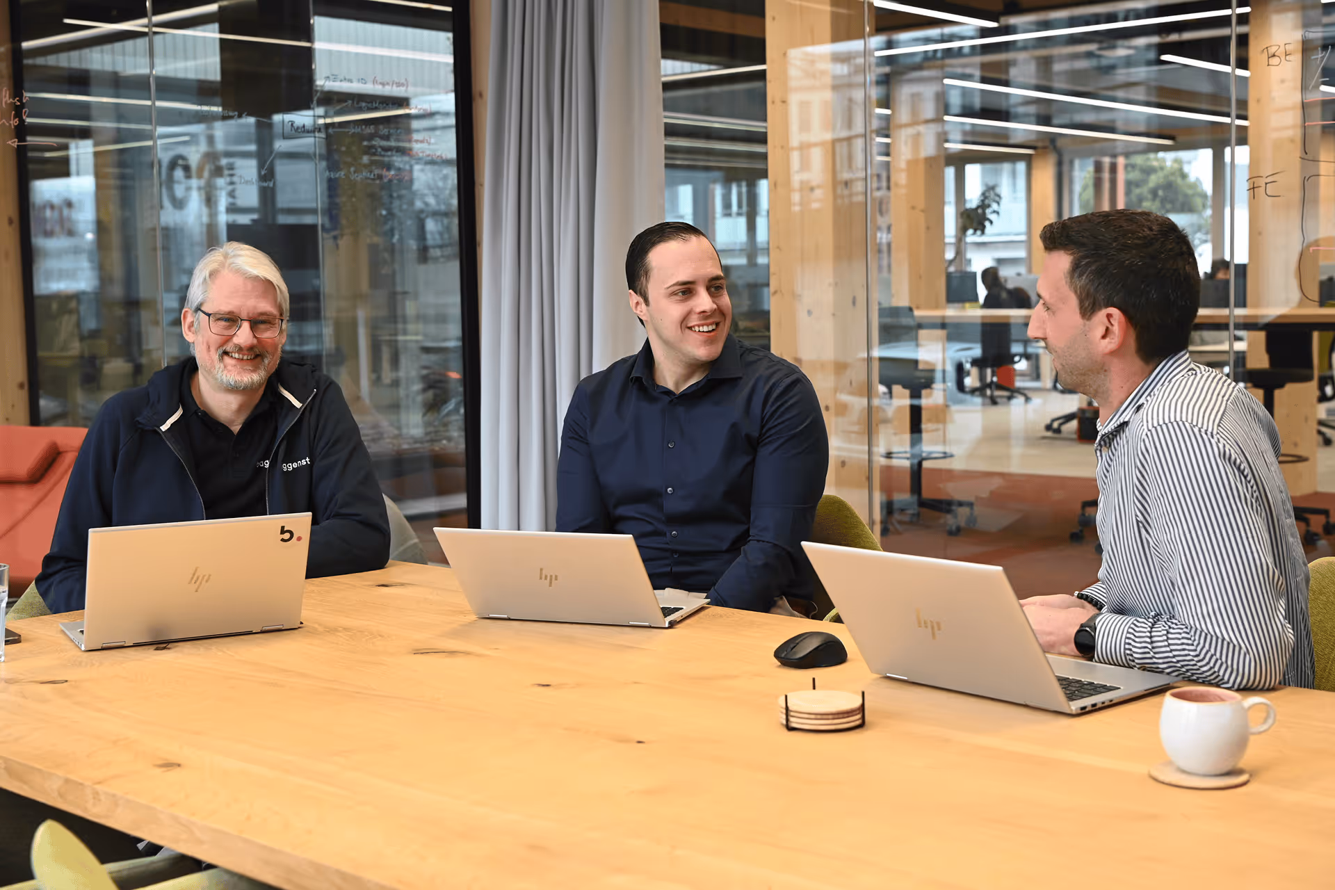 Drei Mitarbeiter mit Laptops an einem Holztisch im modernen Büro